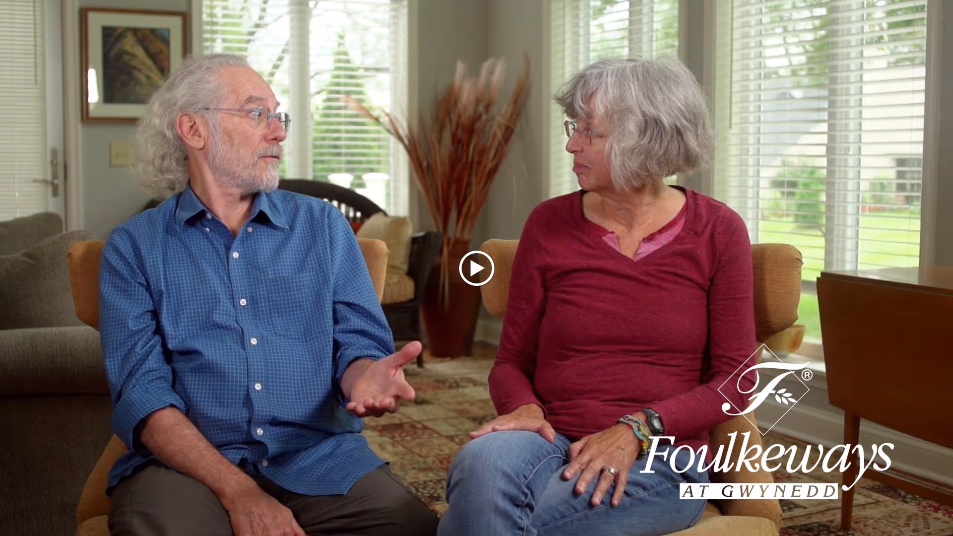 Two older adults seated in a bright living room talking, with large windows and a Foulkeways logo visible.