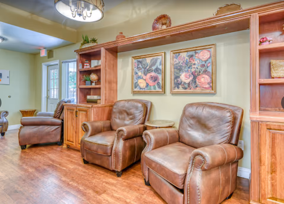 A cozy sitting area with three brown leather armchairs arranged along a wall. Behind the chairs is a wooden built-in shelving unit with decorative items including plants, books, and a globe. Two framed floral paintings hang on the wall above the chairs. The room has wooden flooring and a ceiling light fixture.