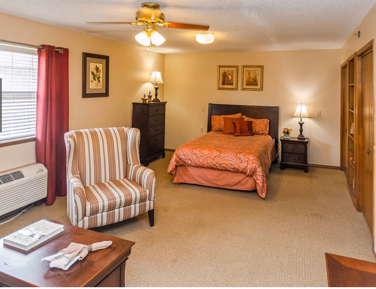 A cozy bedroom with a bed covered in an orange patterned bedspread and multiple pillows. There is a striped armchair near a window with red curtains, a wooden dresser with a lamp and decorative items, a nightstand with a lamp and flowers, and a ceiling fan with lights. The room has beige walls and carpeted floor.