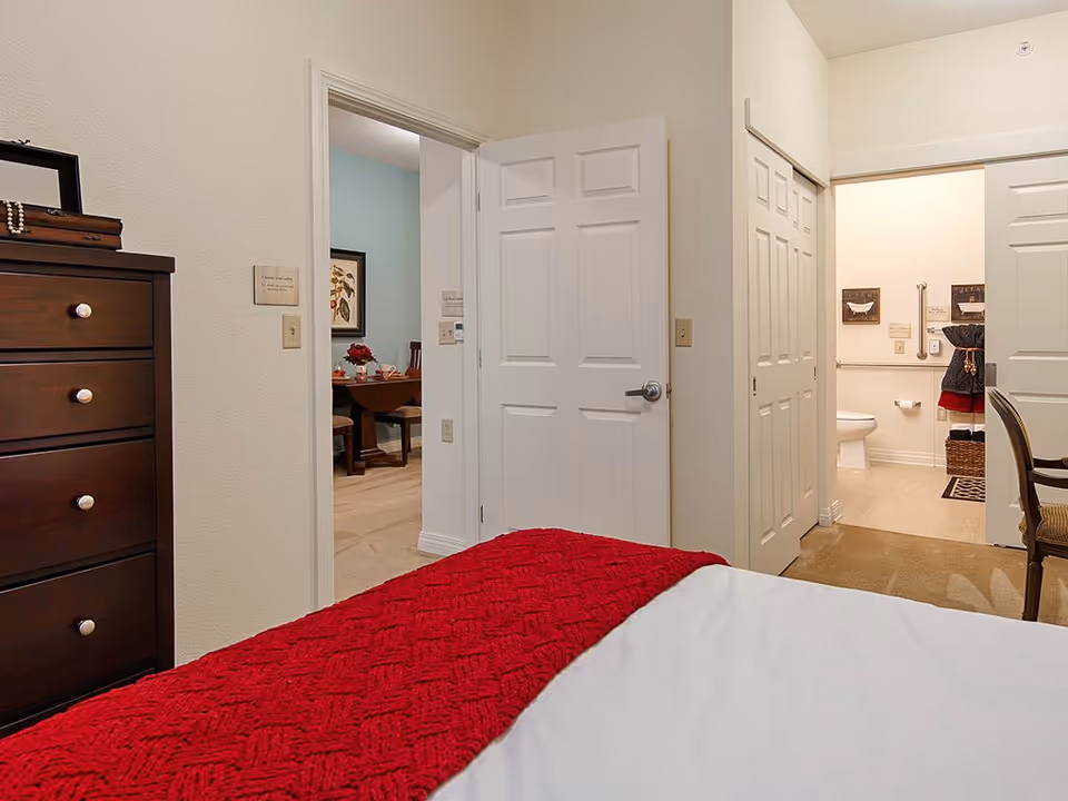 View from a bedroom showing a bed with a red blanket in the foreground, a dark wooden dresser on the left, an open door leading to a dining room with a table and chairs, and another open door revealing a bathroom with a toilet and wall decorations.