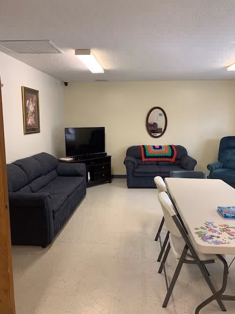 A living room area with two dark blue sofas, one with a colorful crocheted blanket, a flat-screen TV on a black stand, a framed floral painting on the wall, an oval mirror, and a white folding table with chairs around it. There is a puzzle being assembled on the table.