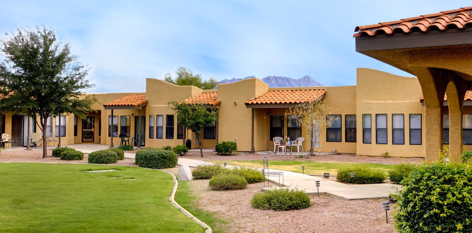 A single-story beige senior living building with red tile roofs surrounding a landscaped courtyard with lawn, walkways, and patio chairs.