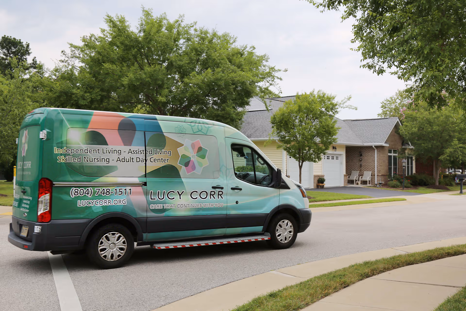 A Lucy Corr facility van parked on a residential street in front of a house with a garage and two chairs on the porch. The van has colorful graphics and text advertising Independent Living, Assisted Living, Skilled Nursing, and Adult Day Center services, along with a phone number and website.