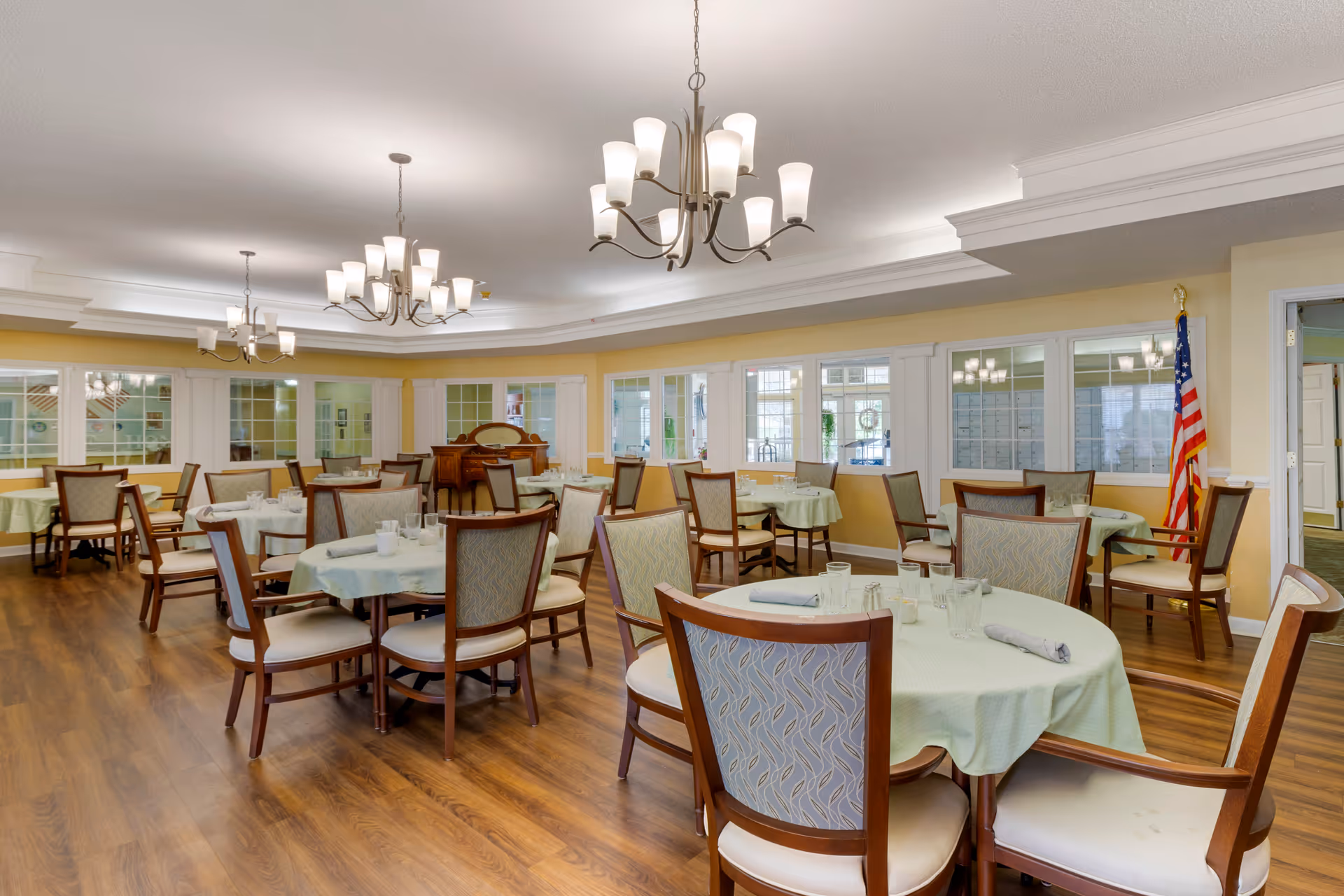 A spacious dining room with multiple round tables covered with light green tablecloths, each set with glasses and napkins. The room has wooden flooring, beige walls, large windows, and several chandeliers hanging from the ceiling. An American flag is visible in the corner.