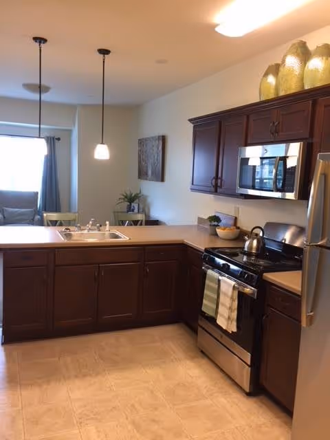 Open kitchen with dark wood cabinets, stainless steel stove and microwave, a sink peninsula, and a view into the adjacent living area.