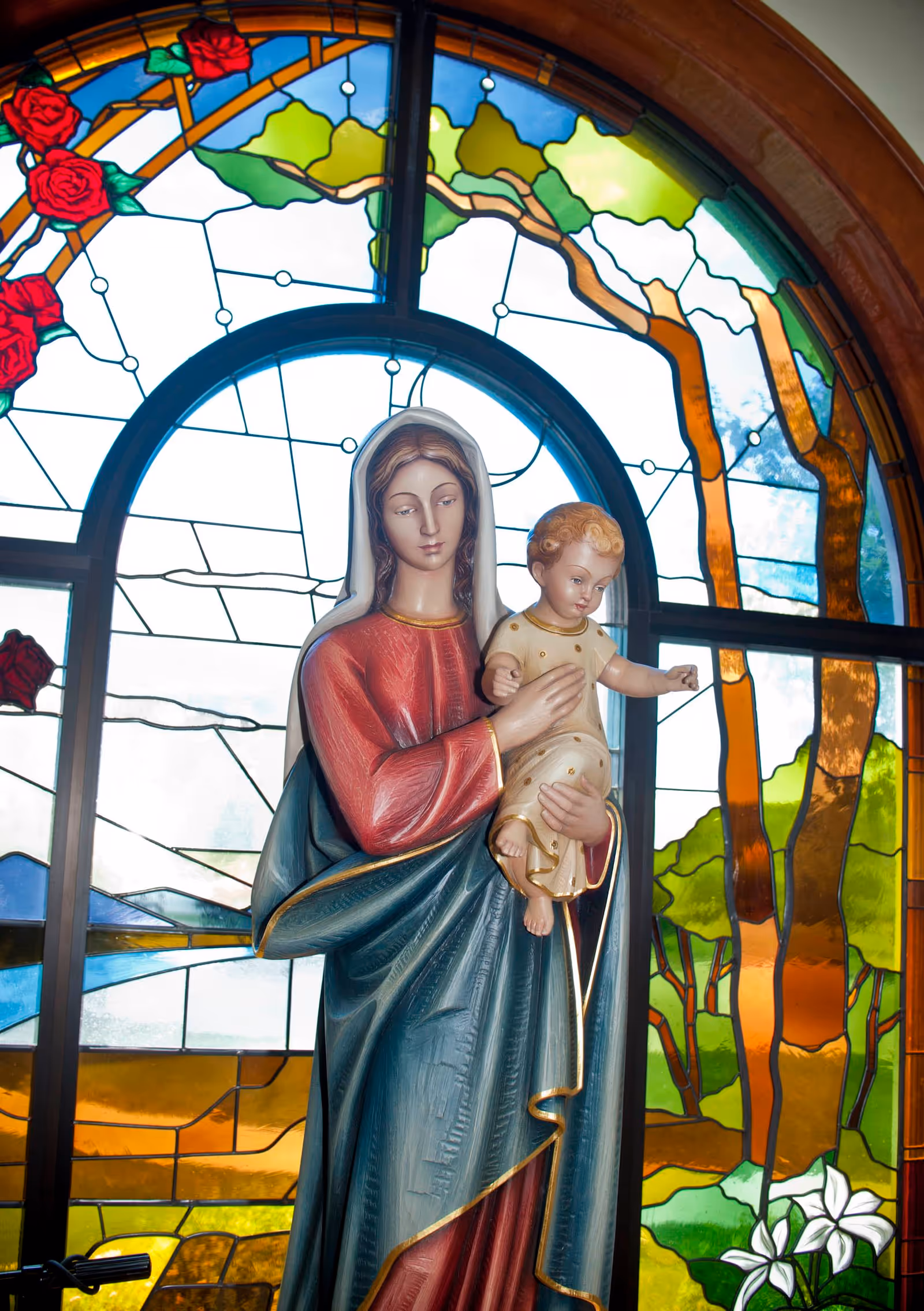 Statue of the Virgin Mary holding baby Jesus in front of a colorful stained glass window depicting trees, flowers, and a scenic landscape.