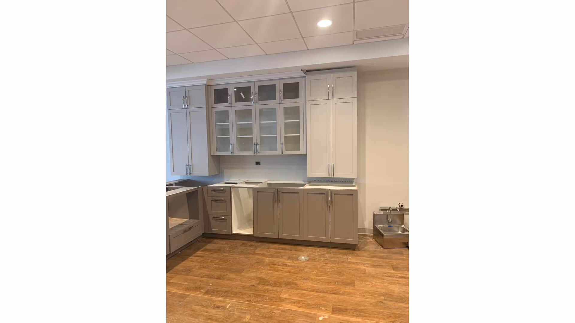 Interior view of a kitchen area with light gray and beige cabinets, some with glass doors, and a wooden floor. The kitchen appears to be in the process of installation or renovation, with some cabinet doors missing and no appliances installed yet. There is a small stainless steel pet water fountain on the floor to the right.