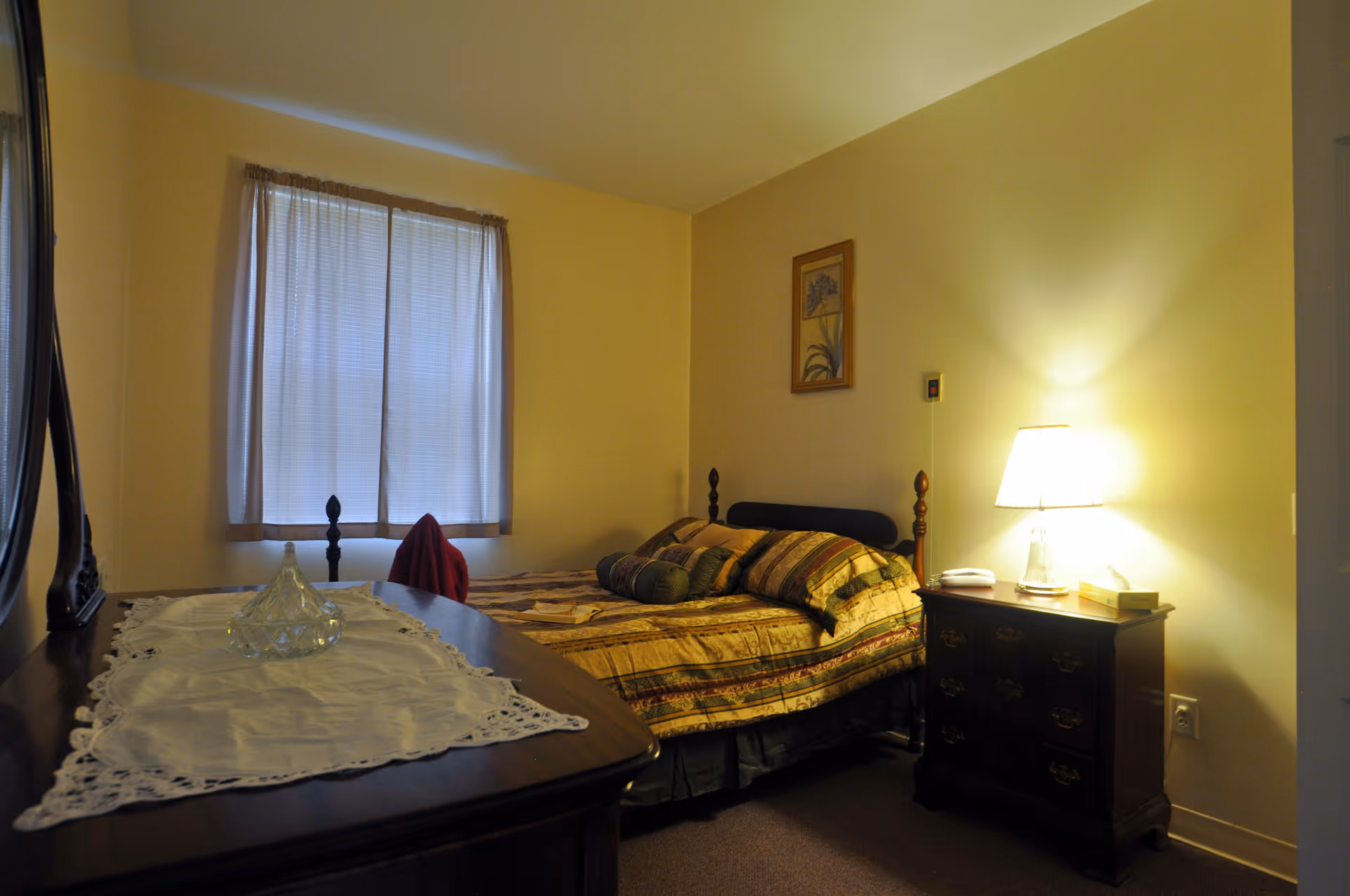 A cozy bedroom with a bed covered in a patterned quilt and multiple pillows. There is a wooden nightstand with a lit lamp, a telephone, and a small box. A framed floral picture hangs on the wall above the bed. A window with sheer curtains allows soft light into the room. A wooden dresser with a lace runner and a glass decorative item is visible in the foreground.