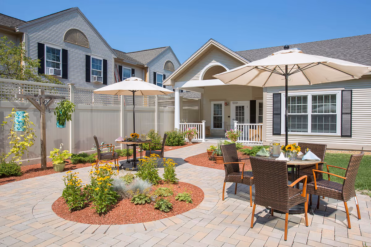 Outdoor patio area at Benchmark Senior Living at Nashua Crossings featuring a paved walkway, garden beds with yellow flowers, and two tables with umbrellas and chairs. The building exterior is visible with beige siding, white trim, and black shutters under a clear blue sky.