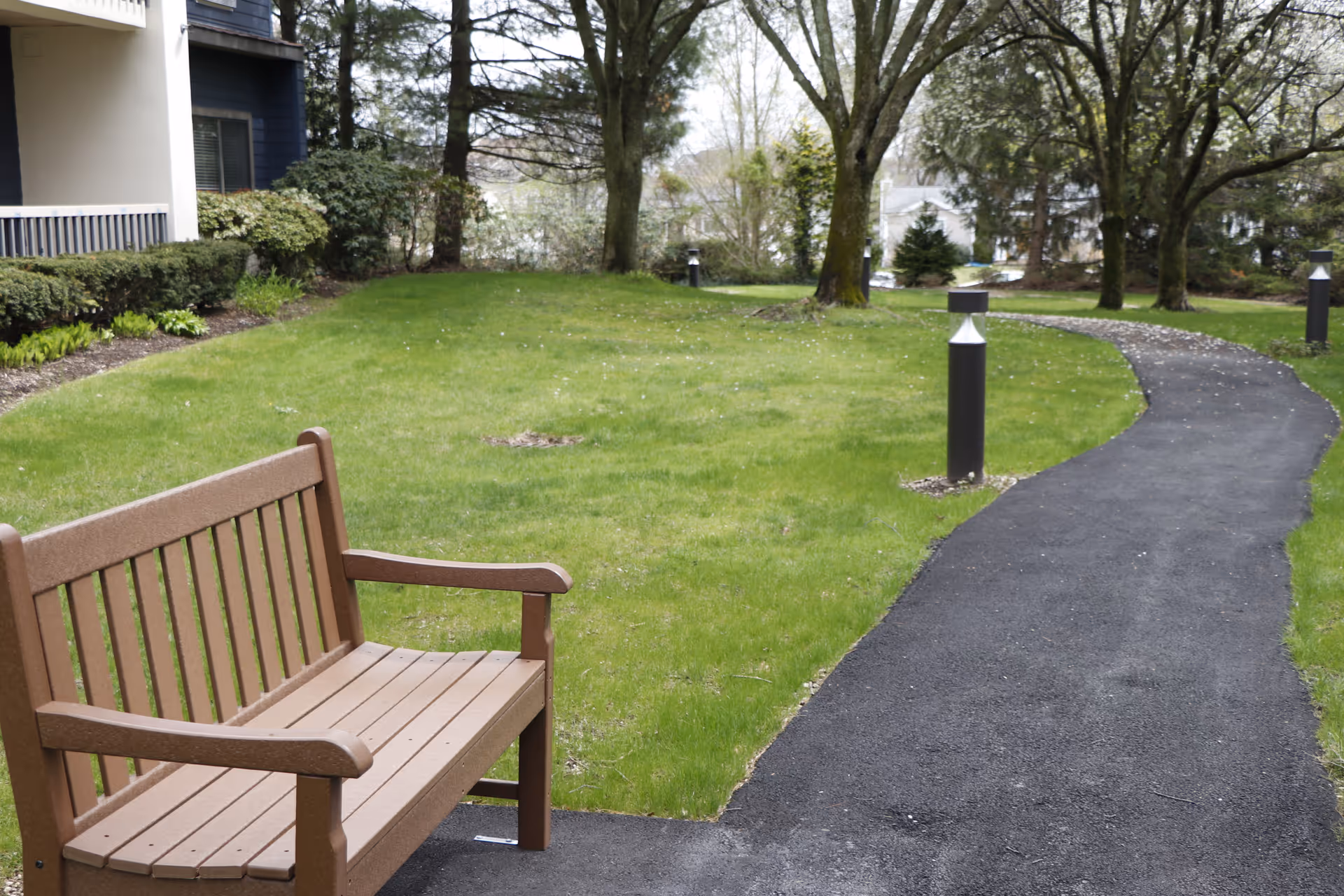 A wooden bench next to a winding paved pathway in a grassy outdoor area with trees and shrubs near a building.