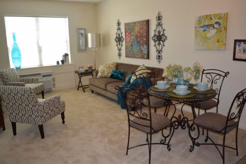 A cozy living room area with a beige carpet, a brown sofa adorned with decorative pillows and a teal throw, two patterned armchairs, a round glass-top dining table with four wrought iron chairs, and several pieces of colorful wall art. A window with blinds lets in natural light, and decorative items including a tall blue vase and small plants are placed on the window sill and side table.