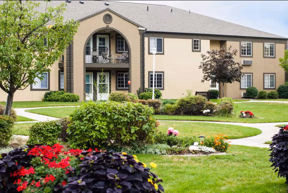 Exterior view of Cedarwood at Sandy, showing a beige two-story building with balconies and multiple windows. The foreground features a well-maintained garden with green bushes, colorful flowers, and a winding concrete pathway.