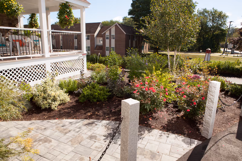 Sunlit outdoor garden and paved walkway in front of a covered porch and brick building.