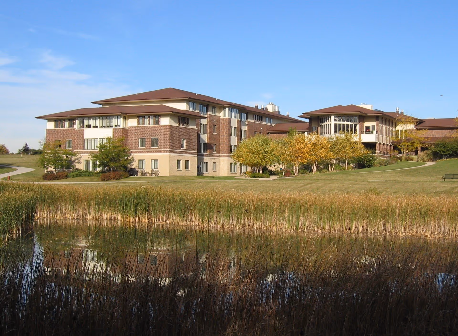A large multi-story senior living facility building with a brown and beige exterior, surrounded by green grass, trees with autumn foliage, and a pond with tall reeds in the foreground reflecting the building.