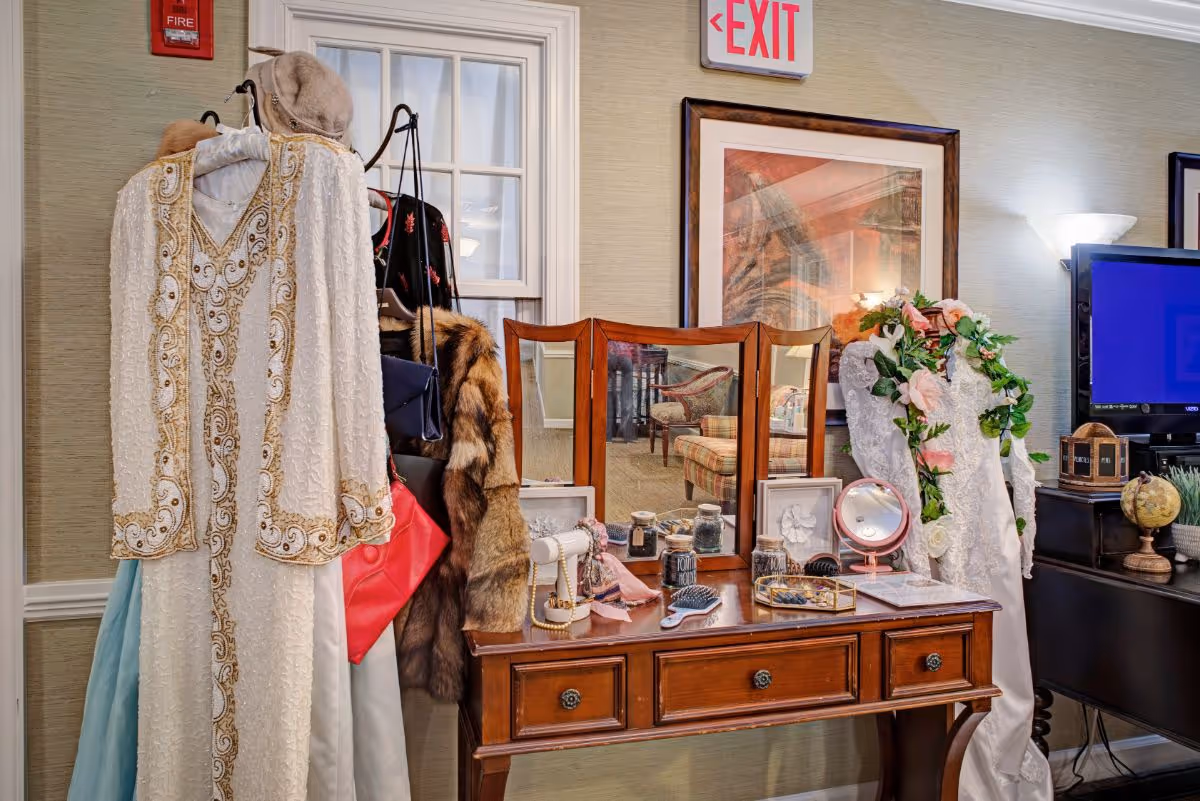 A wooden vanity with a tri-fold mirror displays makeup, brushes and decorative items, flanked by hanging vintage dresses and an EXIT sign on the wall.