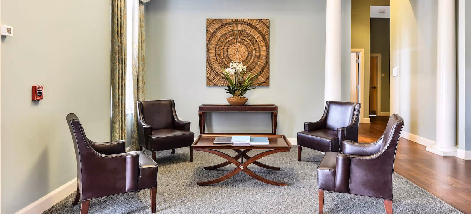 A seating area in a senior living facility with four dark brown leather armchairs arranged around a wooden coffee table. Behind the seating area is a wooden console table with a potted plant and a decorative wall art piece. The room has light-colored walls, a window with curtains, and wooden flooring in the hallway.