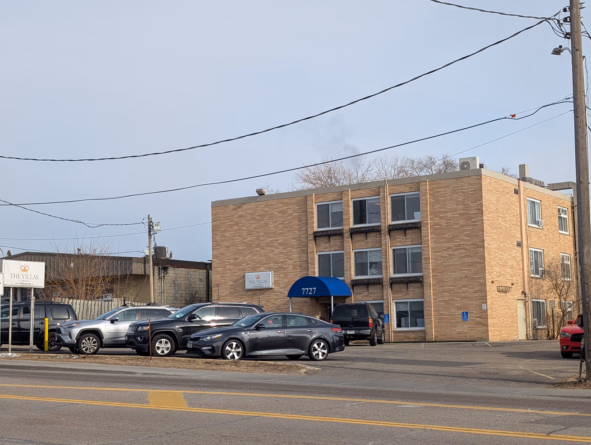 Three-story brick building front with a blue awning over the entrance, parked cars in the lot, and a sign reading 'The Villas'.