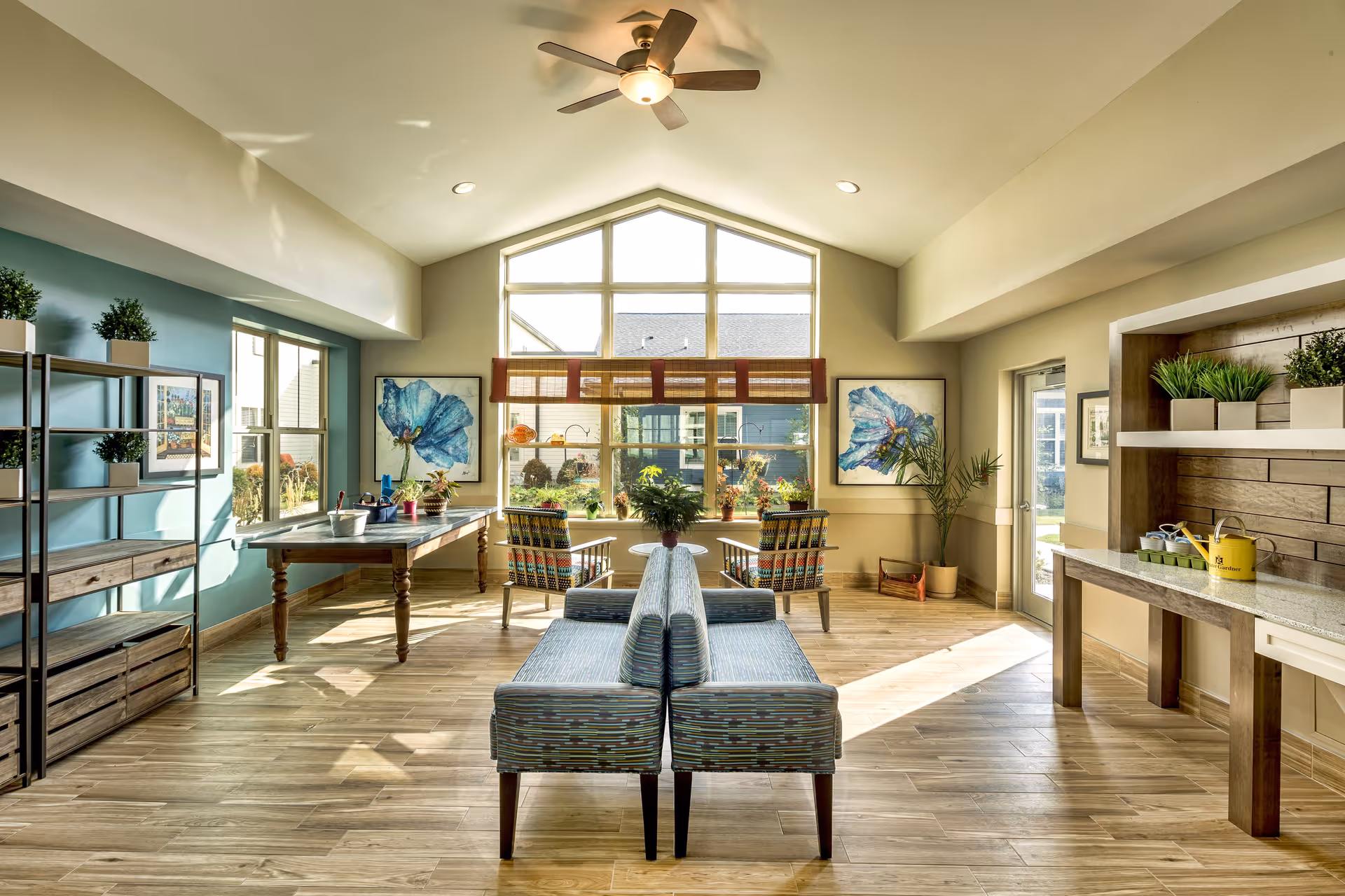 A bright and spacious common area with large windows letting in natural light. The room features two patterned chairs and a small round table near the window, a pair of cushioned benches back-to-back in the center, a wooden table with plants and decorative items on the left, and a countertop with potted plants and a yellow watering can on the right. The walls are painted in soft beige and teal tones, with two large blue floral paintings on either side of the window. The floor is wood, and a ceiling fan is mounted on the high ceiling.