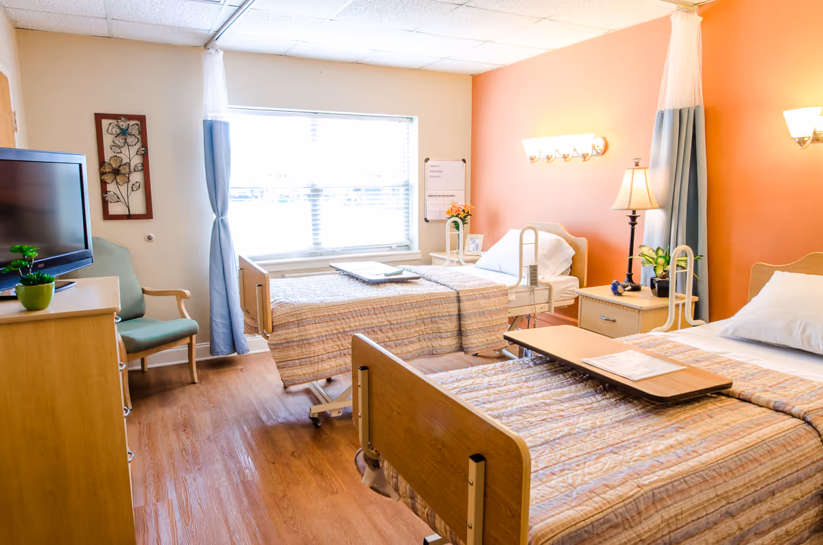 A bright nursing facility room with two adjustable hospital beds, bedside tables, a TV, a chair, and a window.