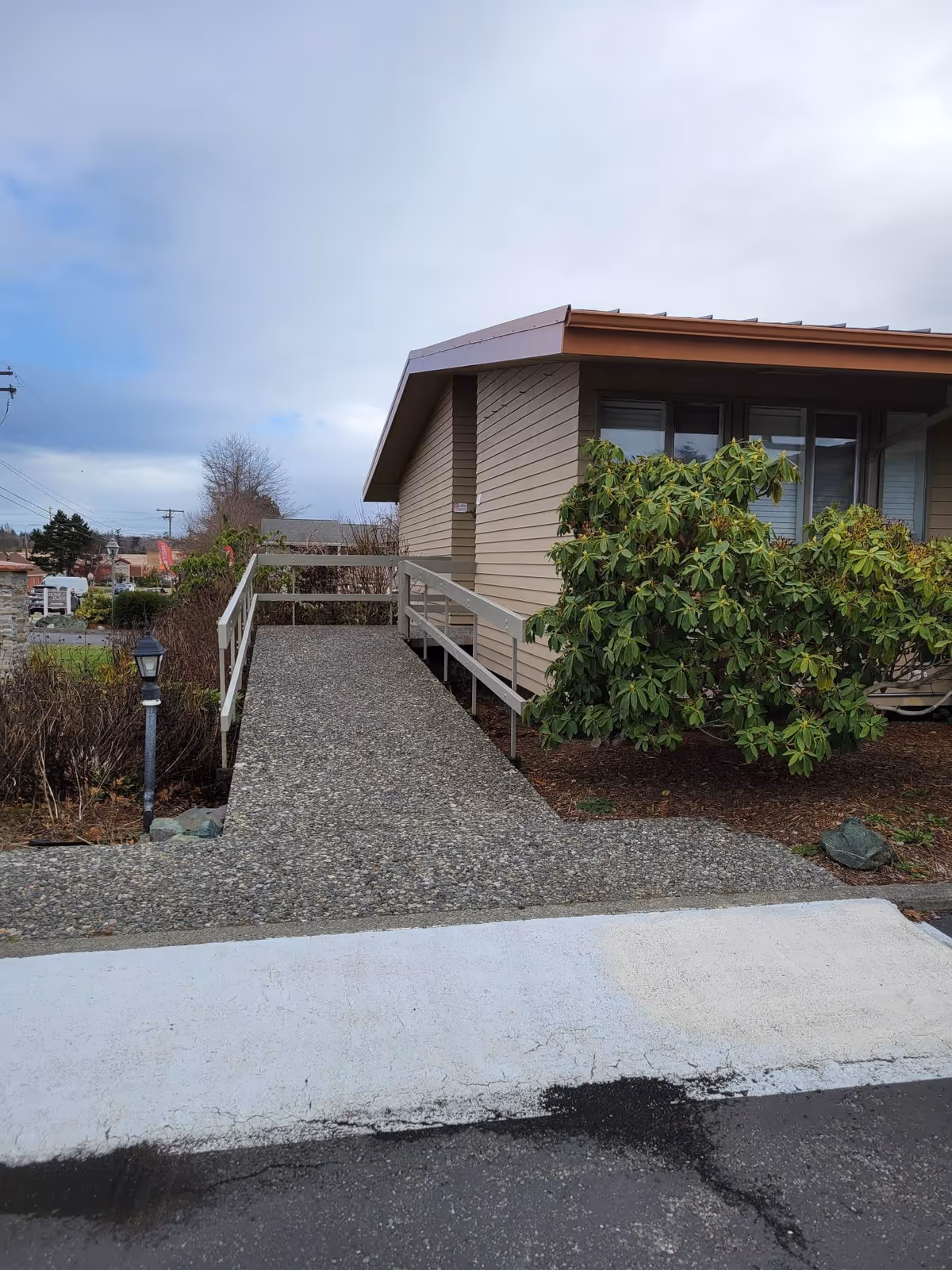 Outdoor view of a beige building with a brown roof and a concrete ramp with metal railings leading up to the entrance. There are bushes and a lamp post on the left side of the ramp, and the sky is cloudy.
