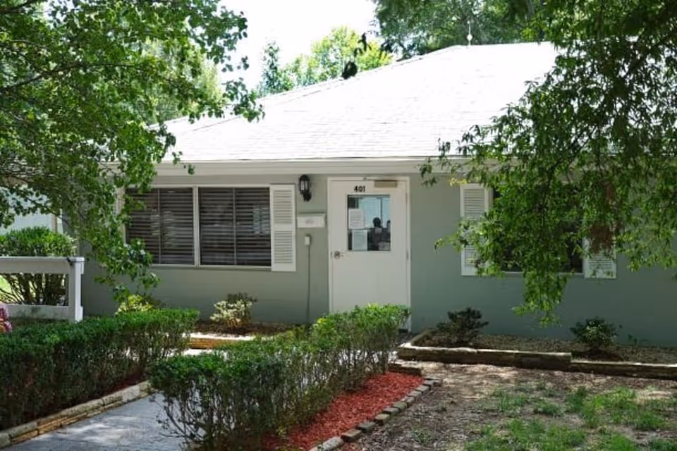 Front exterior view of a single-story building with a white door labeled 401, surrounded by green bushes and trees, with a light green facade and white window shutters.
