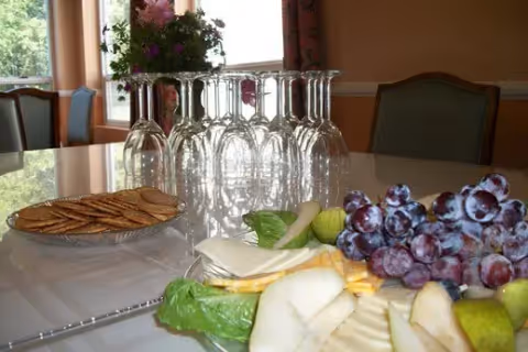 A dining table set with a plate of crackers, a variety of sliced fruits including pears and grapes, and several upside-down wine glasses. In the background, there are chairs and a window with curtains.