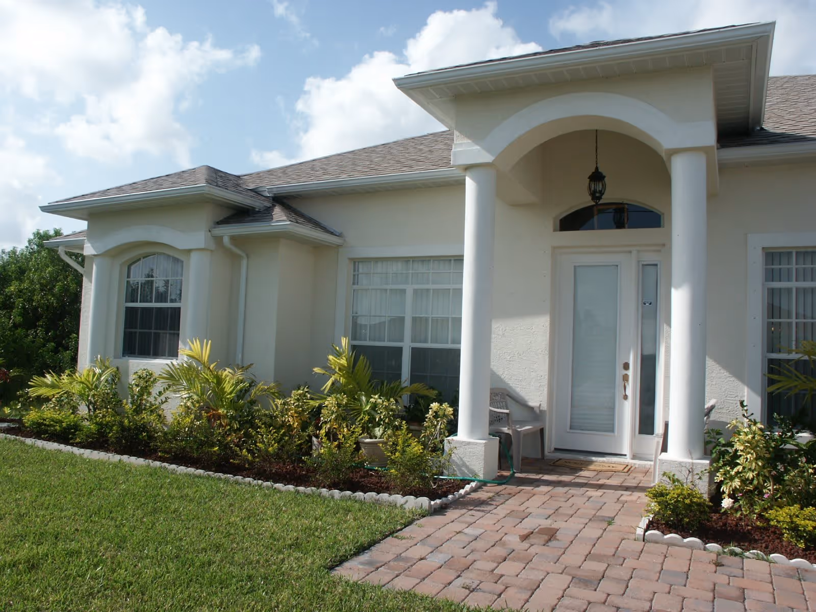 Front exterior view of a single-story building with a light-colored facade, two large windows with white frames, a white front door with glass panels, two white columns supporting a small porch roof, a hanging lantern light fixture, a brick-paved walkway leading to the entrance, and landscaped garden beds with green plants and shrubs on either side of the walkway.