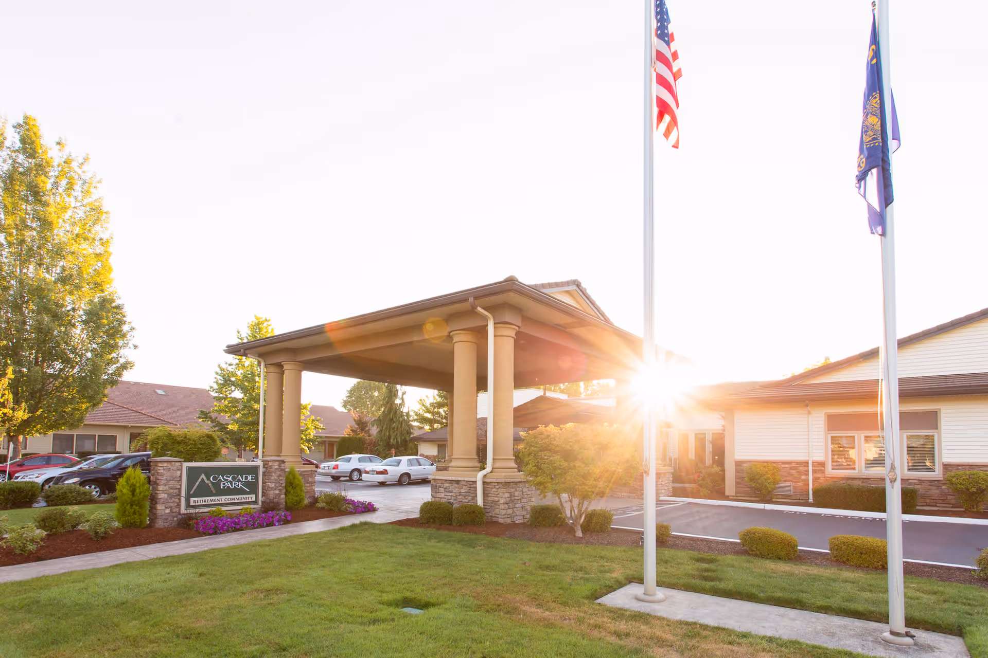Exterior view of Cascade Park Retirement Community entrance with a covered driveway, landscaped greenery, and two flagpoles with the American flag and another flag. The sun is shining brightly in the background.