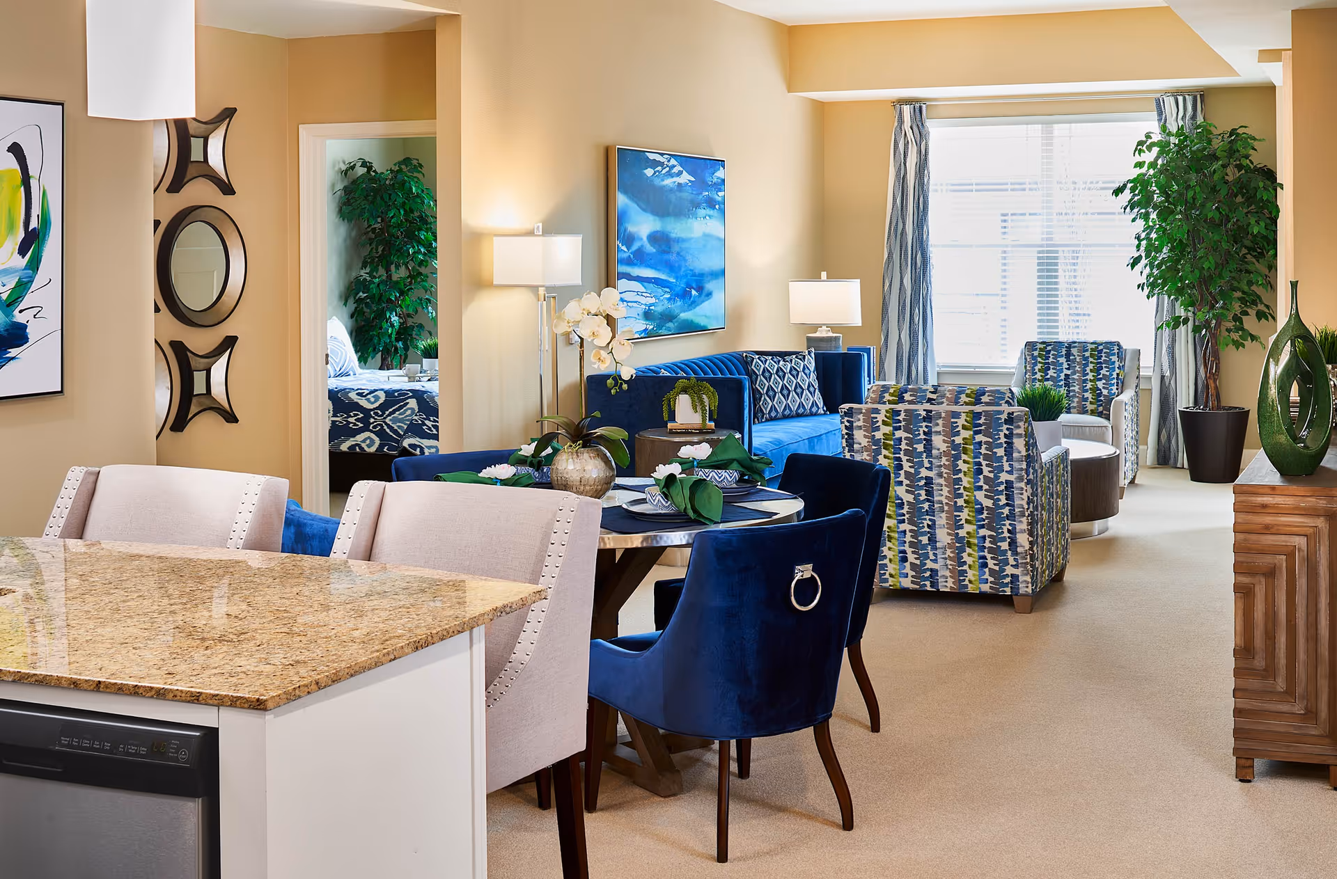 A bright and spacious senior living apartment featuring a granite kitchen island with beige upholstered chairs, a small round dining table with blue velvet chairs, and a living area with a blue sofa, patterned armchairs, and large windows with curtains. The room is decorated with modern artwork, plants, and lamps, creating a cozy and inviting atmosphere. A bedroom is partially visible through an open doorway.