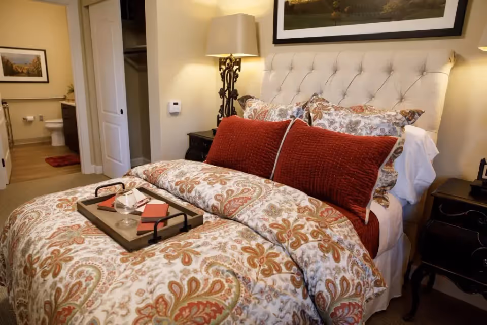 A cozy bedroom with a patterned bedspread and two large red pillows on a tufted headboard bed. A wooden tray with a glass and two red notebooks rests on the bed. There is a bedside table with a decorative lamp and another lamp on the opposite side. In the background, an open door reveals a bathroom with a toilet, a red bath mat, and a framed picture on the wall.