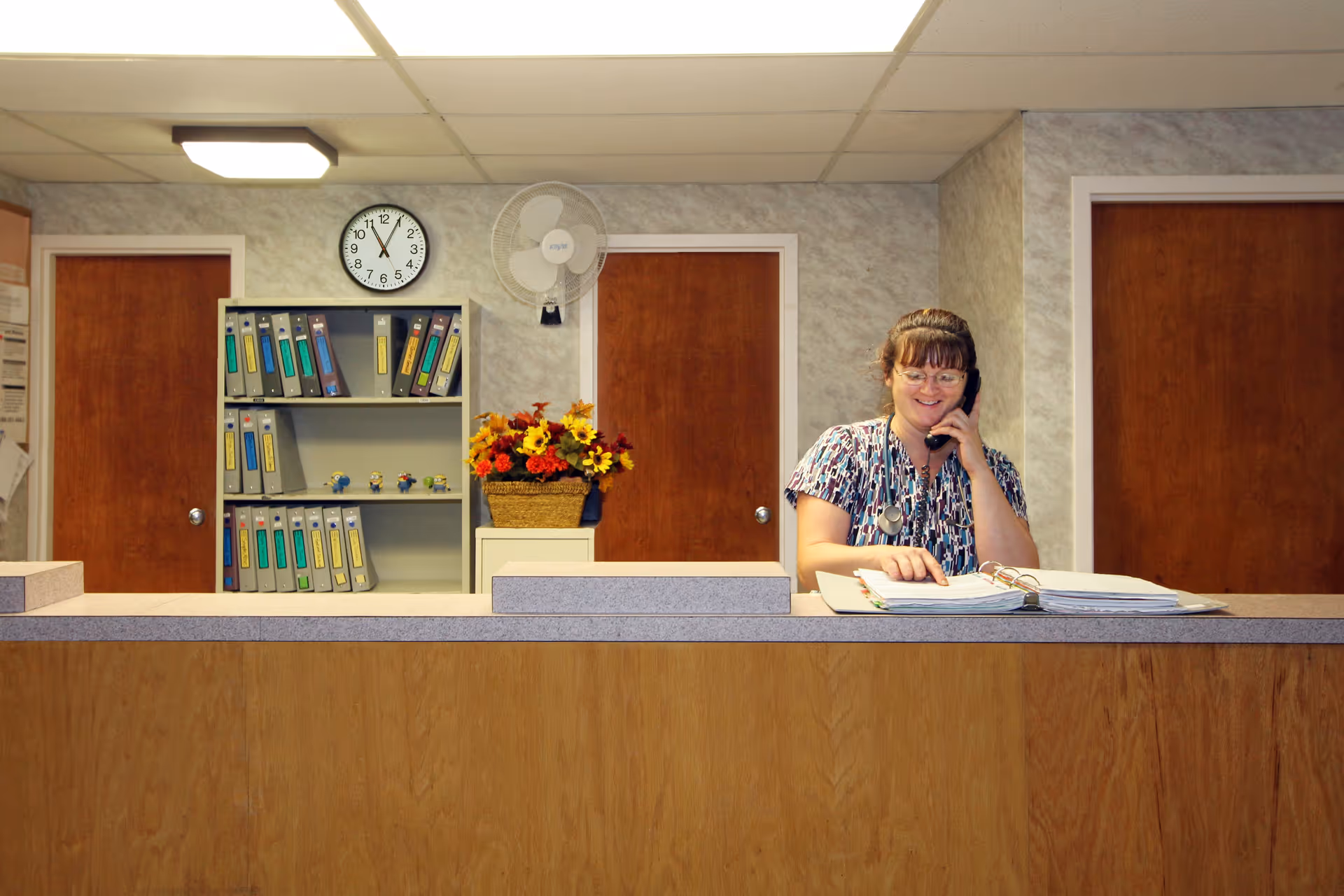 A woman wearing glasses and a patterned scrub top is standing behind a wooden reception desk, talking on a phone and looking at an open binder. Behind her are two closed wooden doors, a wall-mounted fan, a clock showing 11:55, a shelf with binders, and a basket of artificial flowers.