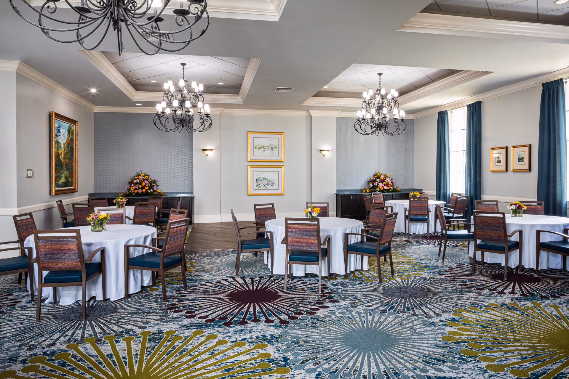 A bright dining/event room with round tables covered in white linens, wooden chairs, chandeliers, and floral centerpieces.