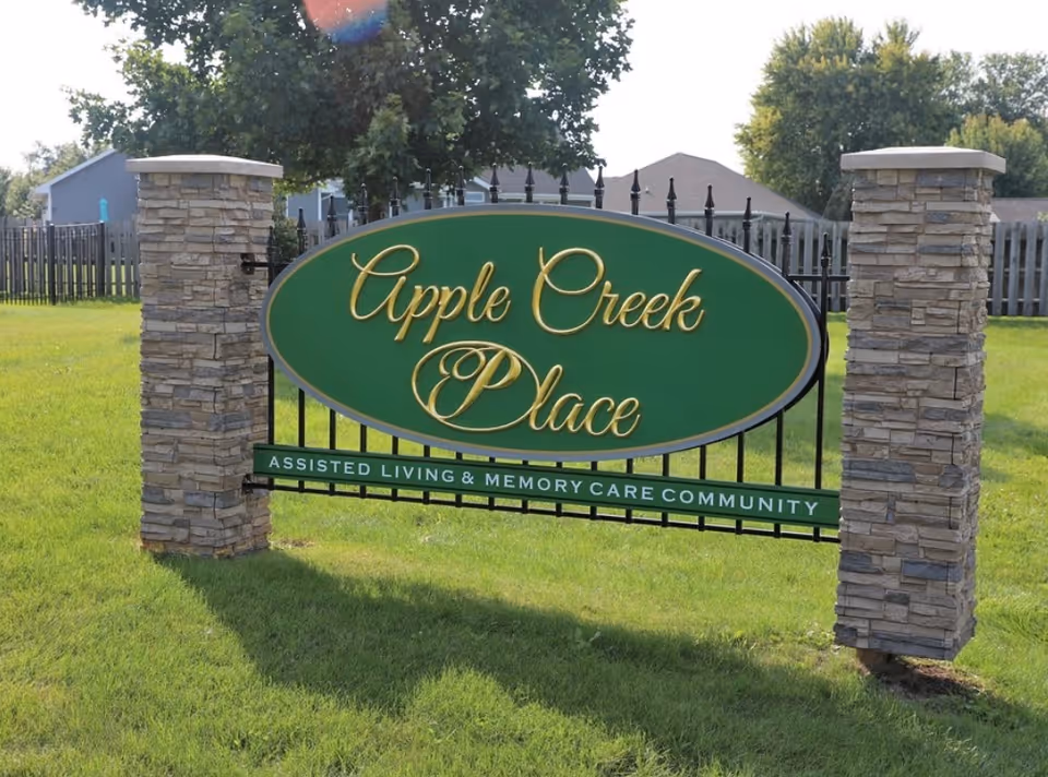 Green oval sign reading "Apple Creek Place" and "Assisted Living & Memory Care Community" mounted between stone pillars on a grassy lawn.