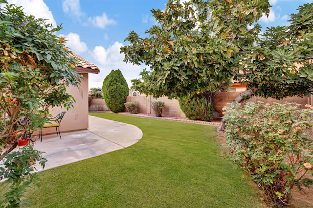 A well-maintained outdoor garden area with green grass, various bushes, and trees. There is a curved concrete pathway leading to a small patio with a chair next to a building with a tiled roof. The sky is blue with some clouds.