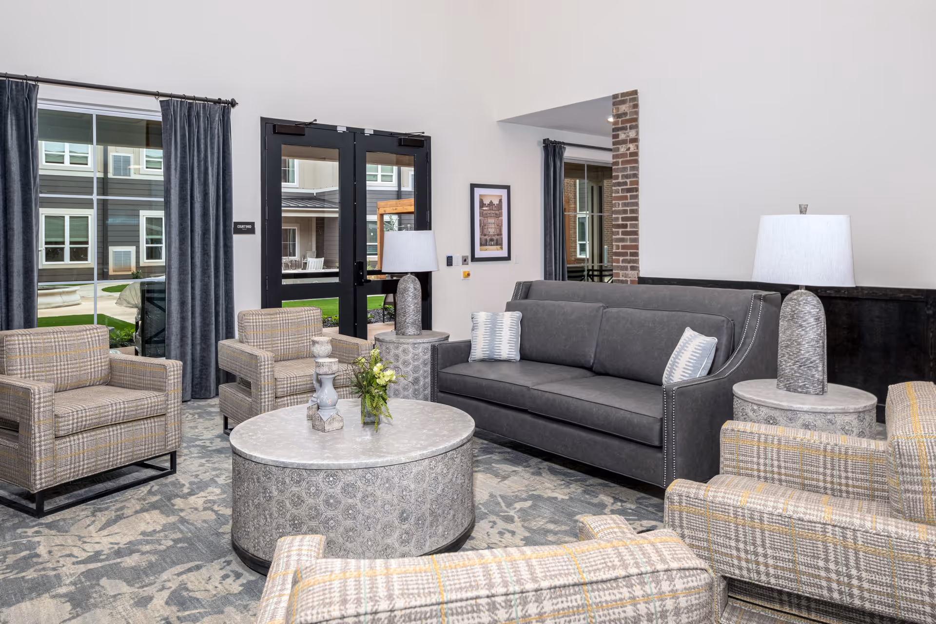 A modern senior living facility lounge area with a gray sofa, four plaid armchairs, two round side tables with lamps, and a round coffee table with decorative items. Large windows and glass doors provide a view of the outdoor courtyard.