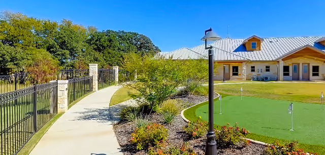 Paved walkway through landscaped grounds with a small putting green and a single-story assisted living building under a clear blue sky.