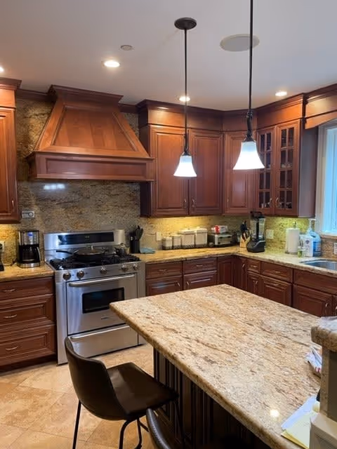 A modern kitchen with dark wooden cabinets, granite countertops, and a central island with a chair. The kitchen features a stainless steel gas stove with an overhead wooden range hood, pendant lights hanging above the island, and various kitchen appliances and containers on the countertops.
