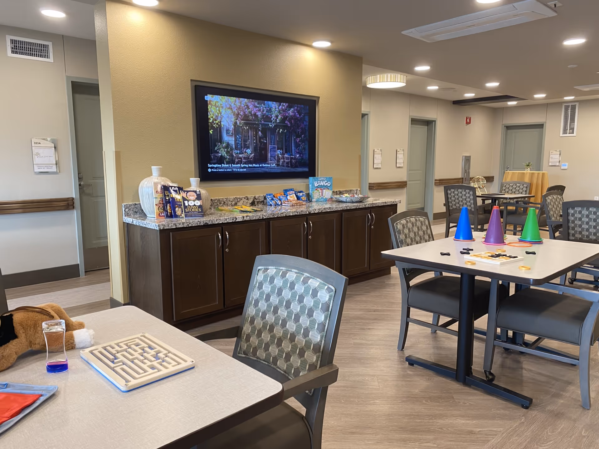 Interior view of a common activity room in a senior living facility with tables and chairs arranged for games and social activities. On one table, there are colorful cone-shaped objects and game pieces, while another table has a maze game and a stuffed animal. A countertop with snacks, books, and a large vase is visible against the wall, above which a flat-screen TV displays an outdoor scene. The room has light-colored walls, wood flooring, and recessed ceiling lights.