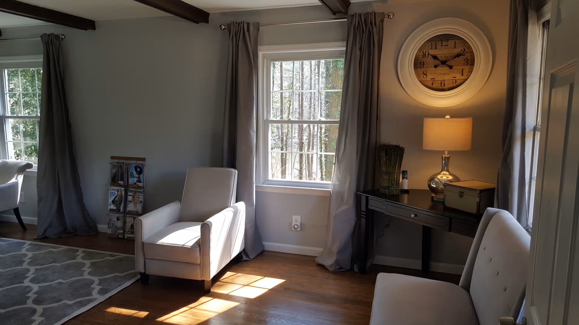 A cozy living room area with two beige armchairs and a beige loveseat. There are two windows with gray curtains letting in natural light. A black console table holds a lamp, a decorative box, and a small plant. A large round wall clock hangs above the table. A magazine rack with several magazines is placed near one of the armchairs. The floor is wooden with a patterned gray and white rug partially visible.