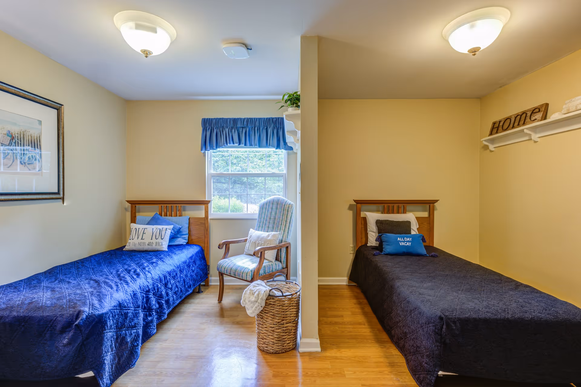 A shared bedroom in a memory care community with two single beds separated by a partial wall. Each bed has a dark blue quilt and decorative pillows. There is a window with a blue valance, a wooden chair with a cushion, a wicker basket, and wall decorations including a framed picture and a shelf with a 'Home' sign.