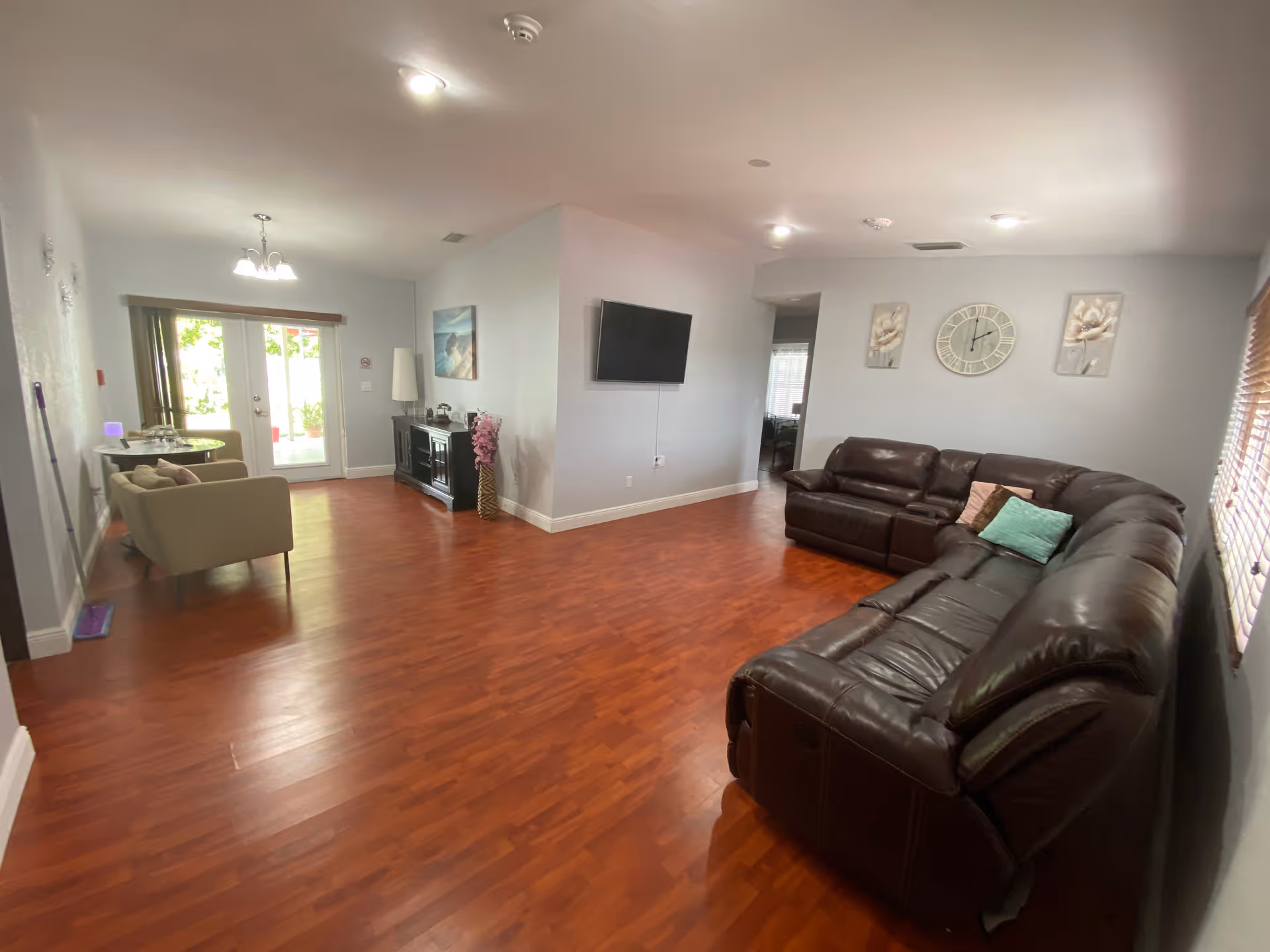 A spacious living room with wooden flooring, featuring a large dark brown leather sectional sofa with colorful pillows on the right side. A flat-screen TV is mounted on a light gray wall in the center. On the left side, there is a small sitting area with two beige armchairs and a round table near glass double doors leading outside. The room is decorated with wall art, a clock, and a tall floor lamp, with natural light coming through the windows with blinds.