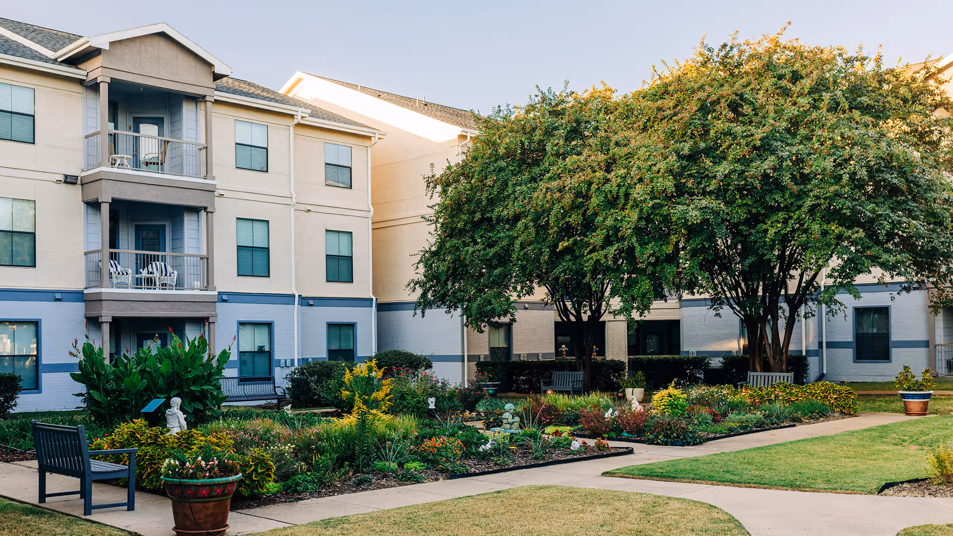 Outdoor garden area with green grass, flower beds, shrubs, and trees in front of a three-story residential building with balconies and windows.
