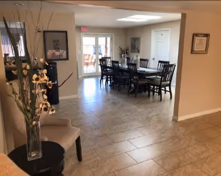 Interior view of a dining area with a long table and chairs, tiled floor, a vase of tall flowers, and glass doors to the outside.
