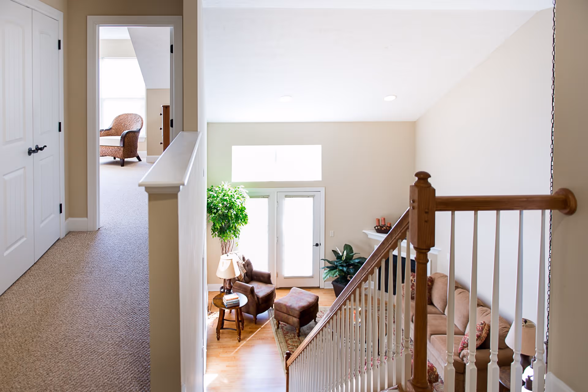 View from the top of a staircase looking down into a living room area with a brown armchair, ottoman, side table with a lamp, and a large potted plant near bright windows. To the left, a hallway leads to another room with a wicker chair and dresser visible.