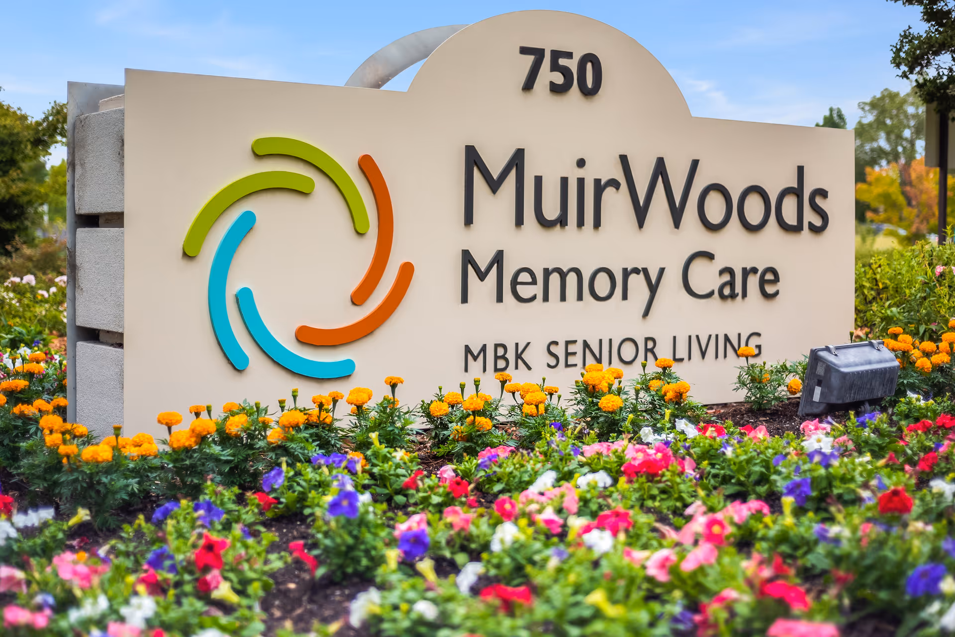 A large beige sign for MuirWoods Memory Care with the address number 750 at the top. The sign features a colorful circular logo with green, orange, and blue segments. The sign is surrounded by a vibrant flower bed with orange, purple, pink, and white flowers, and greenery. The sky is clear and blue in the background.