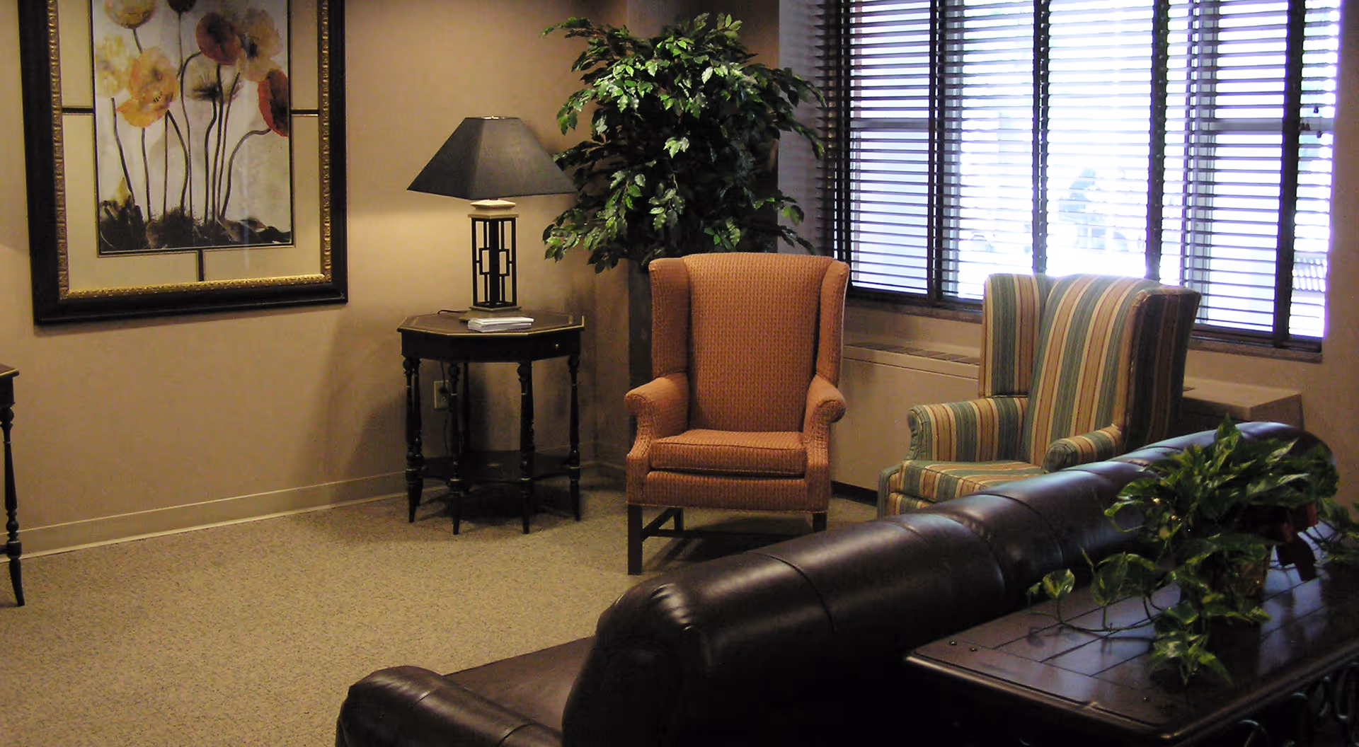 A cozy sitting area with upholstered armchairs, a leather sofa, side table with a lamp, potted plant, and framed artwork by a window with blinds.