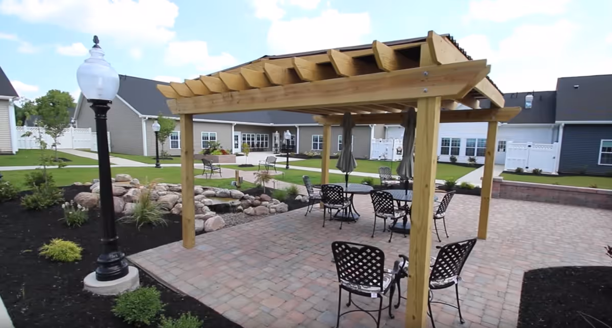Outdoor patio area with a wooden pergola covering several metal tables and chairs. The patio is paved with bricks and surrounded by landscaped garden beds with rocks and plants. In the background, there are single-story buildings with white fences and green lawns under a partly cloudy sky.