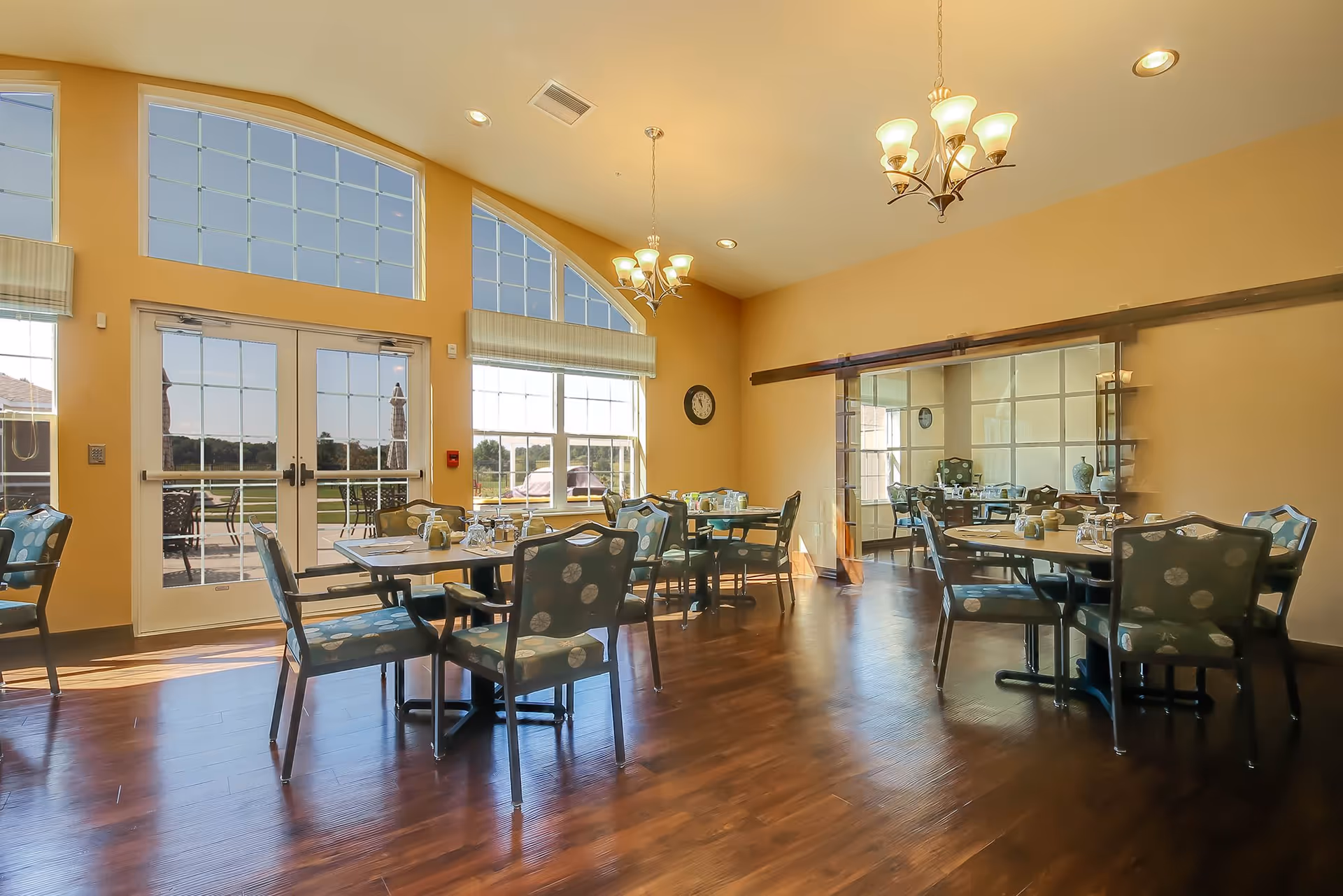 Bright dining room with multiple round tables and chairs, large arched windows and double doors opening to an outdoor patio.