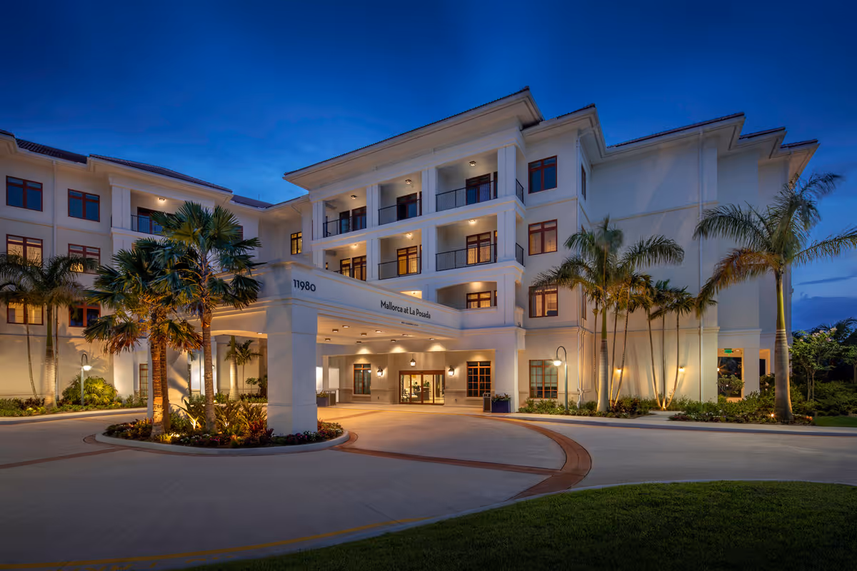 Exterior view of a large, well-lit senior living facility building named Mallorca at La Posada during dusk, featuring palm trees, balconies, and a covered entrance with the address number 11980.