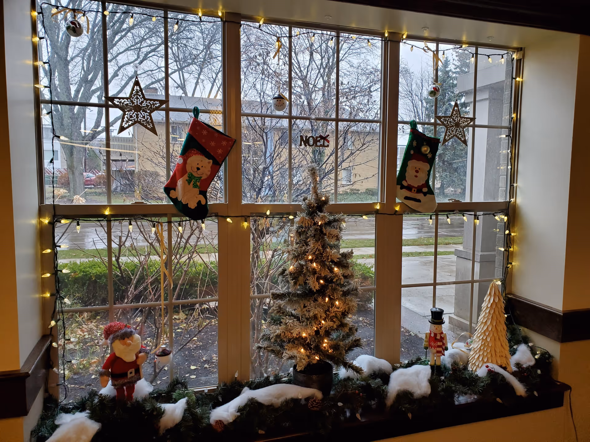 A window decorated with Christmas lights, two hanging stockings featuring a dog and Santa Claus, star ornaments, and a small lit Christmas tree on the windowsill surrounded by festive greenery, cotton snow, a nutcracker figure, and other holiday decorations. Outside the window, bare trees and buildings are visible on a rainy day.